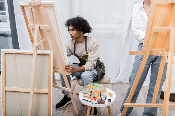 Jovem afro-americano em pintura de avental em tela perto de tintas e namorada em casa — Fotografia de Stock