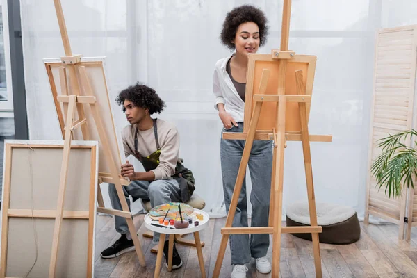 Sonriente mujer afroamericana de pie cerca de lienzo en caballete, novio y pinturas en casa - foto de stock