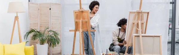 Alegre mujer afroamericana mirando lienzo cerca de novio en casa, bandera - foto de stock