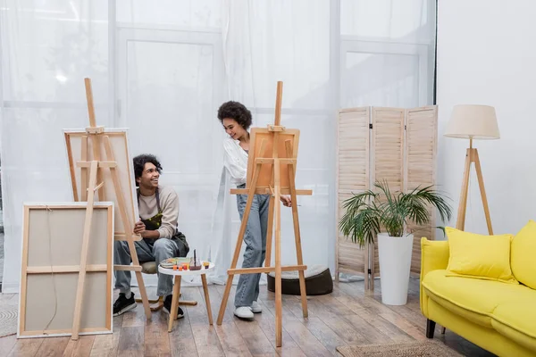 Positiva pareja afroamericana mirándose mientras pintan sobre lienzos en casa - foto de stock