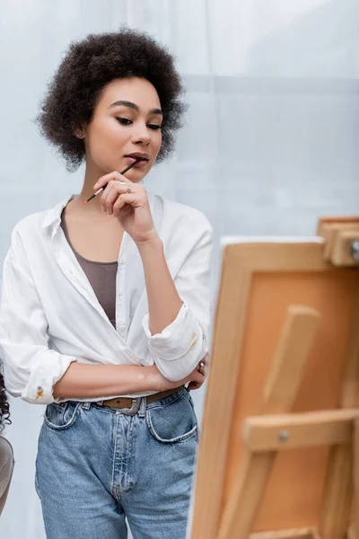 Joven mujer afroamericana sosteniendo pincel cerca de lienzo borroso en casa - foto de stock