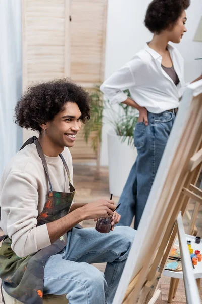 Веселий афроамериканець тримає баночку з фарбами біля полотна і розмиту дівчину вдома. — стокове фото