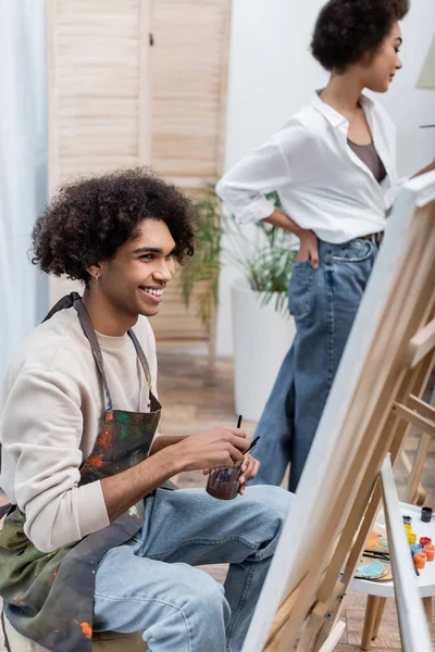 Fröhlicher afrikanisch-amerikanischer Mann hält Glas mit Pinseln in der Nähe von Leinwand und verschwommener Freundin zu Hause — Stockfoto