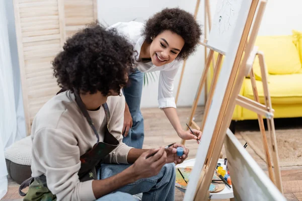 Smiling african american woman holding paintbrush near boyfriend, paints and canvases at home — Stock Photo