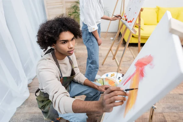 Hombre afroamericano en delantal pintando sobre lienzo borroso cerca de su novia en casa - foto de stock