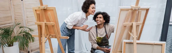 Alegre mujer afroamericana sosteniendo pincel cerca de novio y lienzos en casa, pancarta - foto de stock