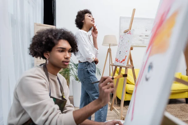 Dreamy african american woman holding paintbrush near canvas and blurred boyfriend painting at home — Stock Photo