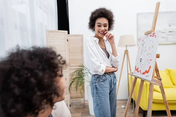 Fröhliche afrikanisch-amerikanische Frau mit Pinsel und Blick auf verschwommenen Freund neben Leinwand auf Staffelei zu Hause — Stockfoto