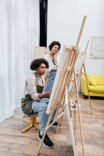 Positive african american woman looking at boyfriend in apron painting on canvas at home — Stock Photo