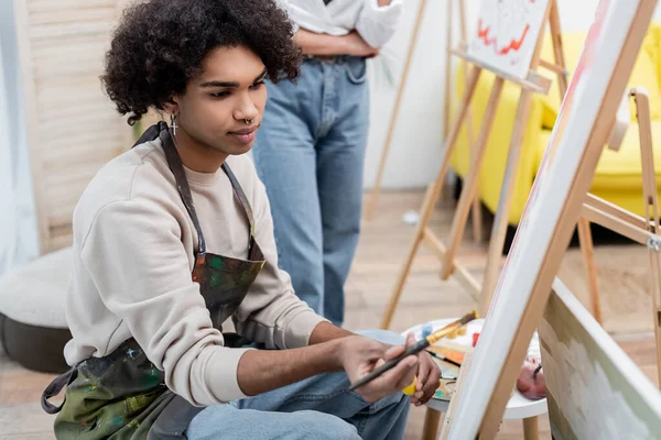 Joven afroamericano hombre pintura sobre lienzo cerca borrosa novia en casa - foto de stock
