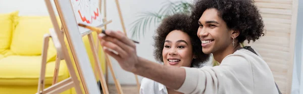 Fröhliche afrikanisch-amerikanische Mann Malerei auf Leinwand in der Nähe junge Freundin zu Hause, Banner — Stockfoto