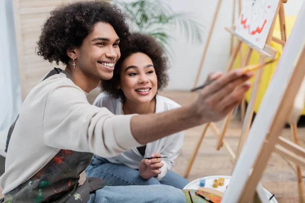 Jeune homme afro-américain peignant sur toile floue sur chevalet près de petite amie avec pinceau à la maison — Photo de stock