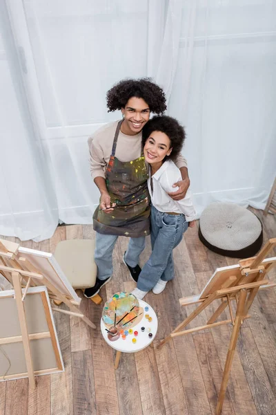 Вид сверху на улыбающуюся афроамериканскую пару, смотрящую в камеру возле красок и мольбертов дома — стоковое фото