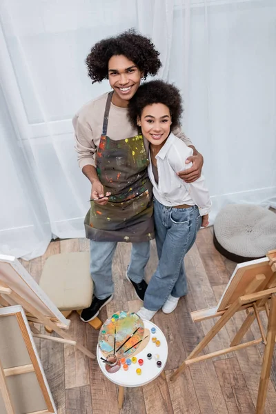 Draufsicht auf fröhliche afrikanisch-amerikanische Mann umarmt Freundin in der Nähe von Staffeleien und Leinwänden zu Hause — Stockfoto