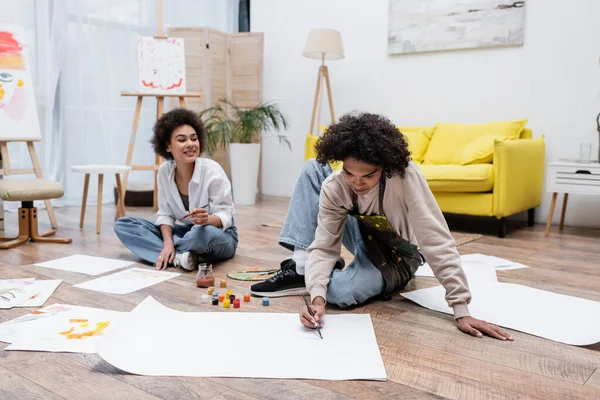 African american man painting on floor near smiling girlfriend and paints at home — Stock Photo