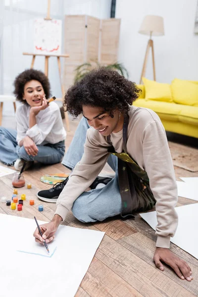 Sorridente uomo africano americano pittura sul pavimento vicino fidanzata offuscata a casa — Foto stock