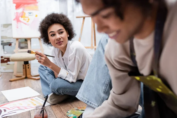 Young african american woman holding paintbrush near blurred boyfriend and papers on floor — Stock Photo