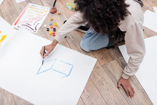 Vista aérea de la pintura de hombre afroamericano sobre papel cerca de pinturas en el suelo en casa - foto de stock