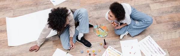 Vue aérienne d'une femme afro-américaine joyeuse regardant son petit ami peindre sur le sol à la maison, bannière — Photo de stock