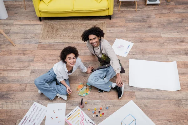 Top view of cheerful african american couple looking at camera near papers and paints on floor — Stock Photo