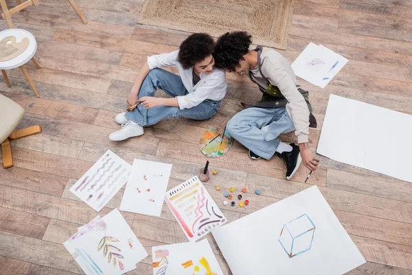 Overhead view of cheerful african american couple holding paintbrushes near papers and paints on floor — Stock Photo