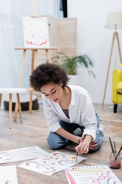 Africano mulher americana pintura em papel perto pincéis em frasco no chão em casa — Fotografia de Stock