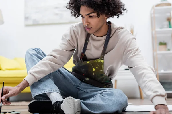 African american man in apron holding paintbrush while sitting on floor at home — Stock Photo