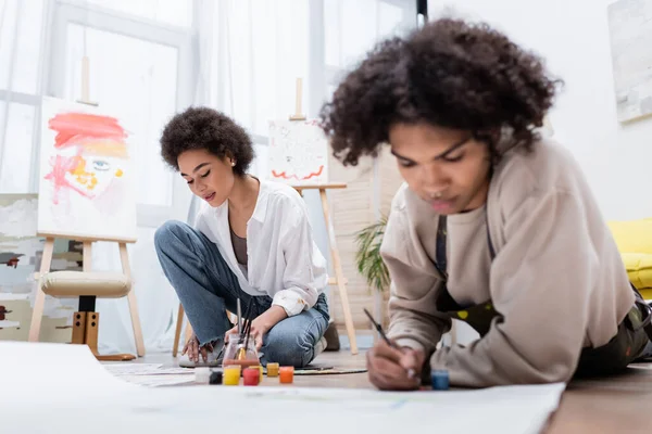 African american woman painting on paper near blurred boyfriend on floor in living room — Stock Photo
