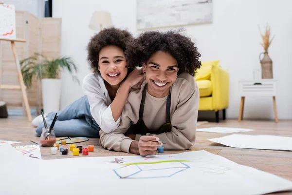 Happy african american woman hugging boyfriend and looking at camera near papers and paints on floor — Stock Photo