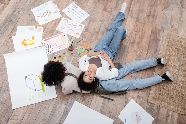 Top view of african american woman smiling at camera while lying near boyfriend painting on floor — Stock Photo