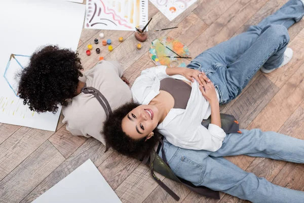 Blick von oben auf eine afrikanisch-amerikanische Frau, die in die Kamera lächelt und in der Nähe ihres Freundes auf dem Boden im Wohnzimmer malt — Stockfoto