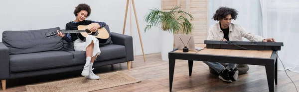 Mulher afro-americana alegre tocando guitarra acústica perto do namorado com sintetizador em casa, banner — Fotografia de Stock