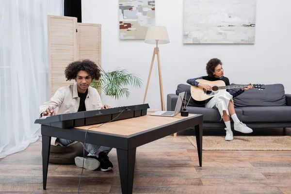 Joven afroamericano jugando sintetizador cerca de la computadora portátil y novia borrosa con guitarra acústica - foto de stock