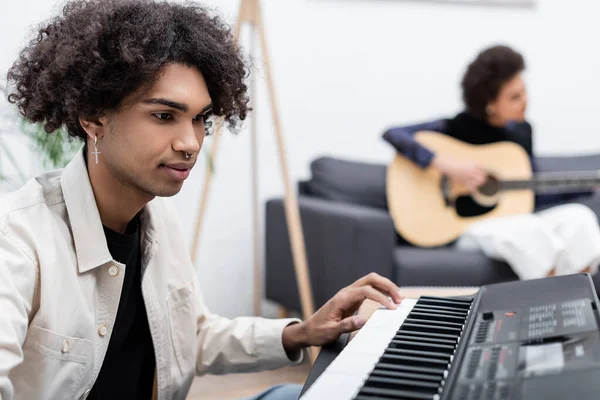 Jovem afro-americano tocando sintetizador perto de namorada borrada com guitarra acústica em casa — Fotografia de Stock