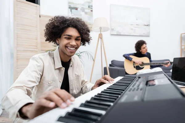 Sorridente uomo africano americano che gioca sintetizzatore vicino al computer portatile e fidanzata offuscata con chitarra acustica a casa — Foto stock