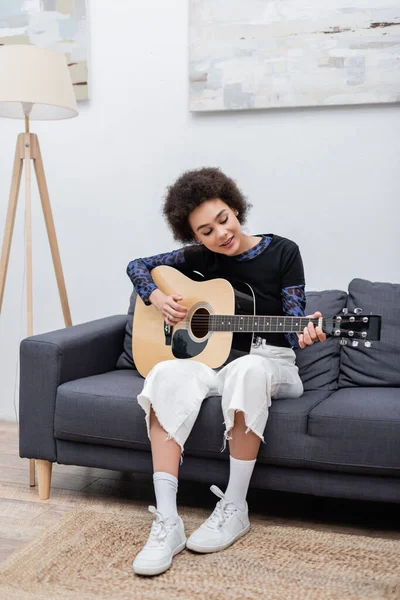 Sonriente mujer afroamericana tocando la guitarra acústica en el sofá en casa - foto de stock