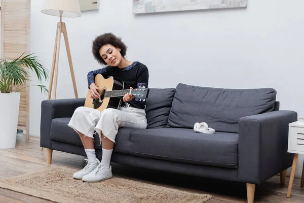 Mujer afroamericana joven tocando la guitarra acústica cerca de los auriculares en el sofá - foto de stock