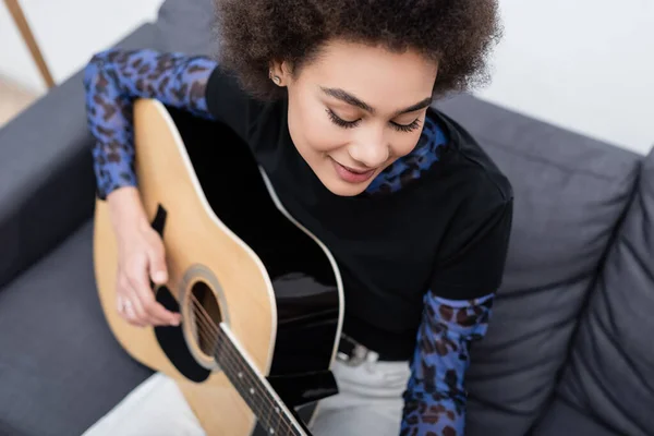 Blick aus der Vogelperspektive auf eine lächelnde Afroamerikanerin, die zu Hause unscharfe Akustikgitarre spielt — Stockfoto