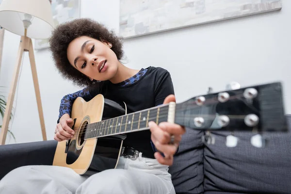 Mujer afroamericana joven tocando la guitarra acústica en el sofá en la sala de estar - foto de stock
