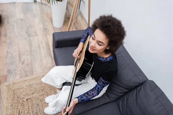 Vista aérea de la mujer afroamericana positiva tocando la guitarra acústica en el sofá en casa - foto de stock