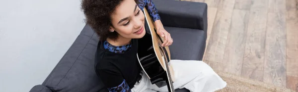 Vue aérienne de la femme afro-américaine jouant de la guitare acoustique sur le canapé dans le salon, bannière — Photo de stock