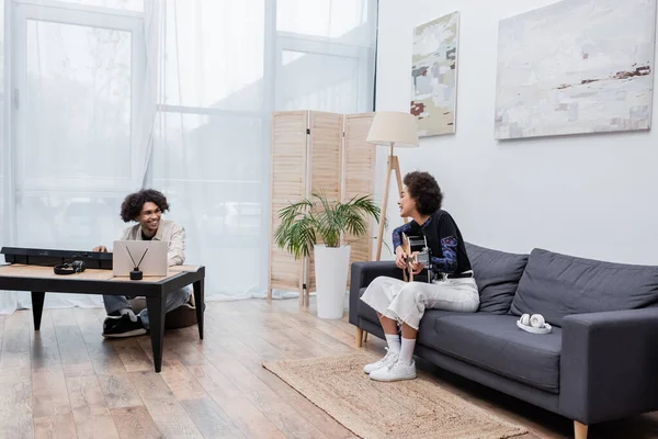 Sonriente pareja afroamericana tocando instrumentos musicales cerca de auriculares y laptop en casa - foto de stock