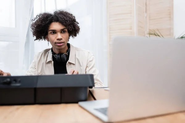 Uomo afroamericano con le cuffie guardando il computer portatile offuscato vicino al sintetizzatore a casa — Foto stock