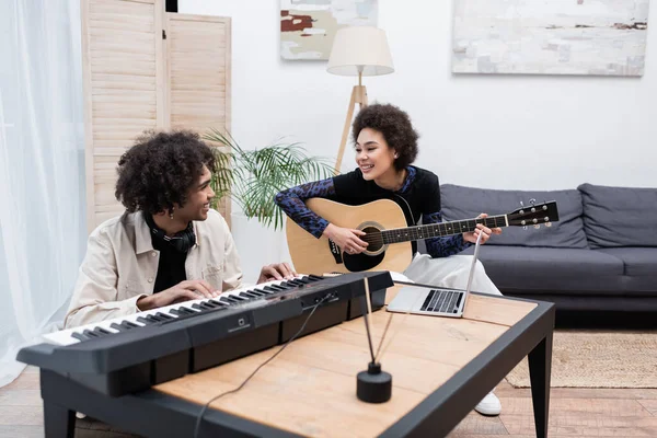 Mulher afro-americana alegre tocando guitarra acústica perto do namorado com sintetizador em casa — Fotografia de Stock