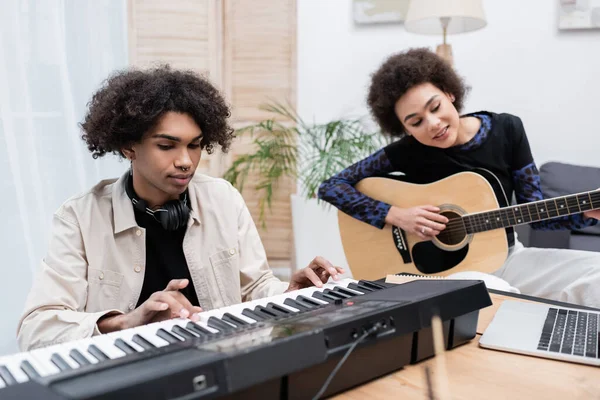 Uomo afroamericano che gioca sintetizzatore vicino sorridente fidanzata con chitarra acustica a casa — Foto stock