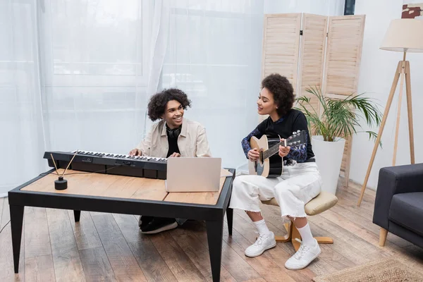 Smiling african american woman playing acoustic guitar near boyfriend with synthesizer and laptop in living room — Stock Photo