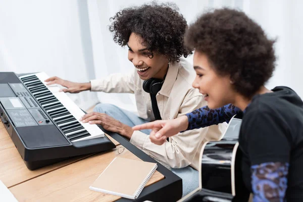 Smiling african american man playing synthesizer near girlfriend with acoustic guitar at home — Stock Photo