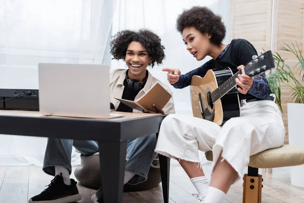 Sorrindo homem afro-americano olhando para laptop borrado perto de sintetizador e namorada com guitarra acústica em casa — Fotografia de Stock