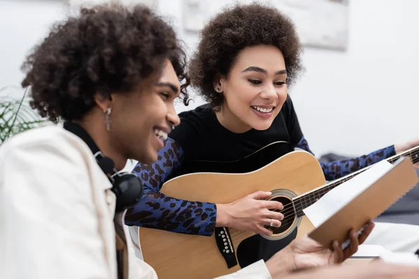 Jovem afro-americano segurando notebook perto namorada tocando guitarra acústica em casa — Fotografia de Stock