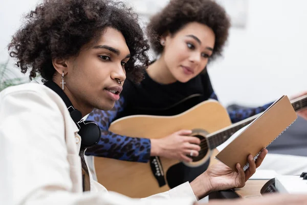 Africano americano com fones de ouvido segurando notebook perto namorada borrada tocando guitarra acústica e sintetizador em casa — Fotografia de Stock